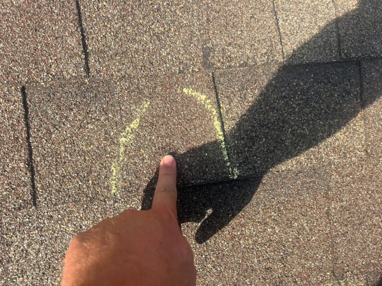 Close-up of hail damage on an asphalt roof shingle with a finger pointing at the impact mark.